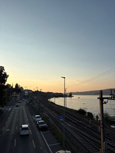 The image depicts a sunset view of the city of Wädenswil, Switzerland, featuring a prominent lake, railway tracks, and a road. In the foreground, a road with lane markings is visible, with several cars driving and parked. A white hatchback is in the left lane, followed by a dark-colored sedan in the right lane. Further down the road, a white SUV and a dark sedan are parked on the right side. To the right of the road, an extensive network of railway tracks runs parallel to the water. A blue parking sign is visible near the tracks.
The lake, likely Lake Zurich, occupies the middle and right portions of the image, reflecting the warm hues of the sunset. A small boat is visible on the water. The horizon is marked by a distant shoreline with hills. On the left side of the image, buildings characteristic of a town or city line the road, including houses and a modern building with the name "wabi" visible. A church spire can be seen among the buildings. Trees are present on the far left, casting shadows. The sky is a gradient of light blue and soft orange, indicative of dusk. Electrical poles and wires are present, particularly near the railway tracks. The scene is captured from an elevated perspective.