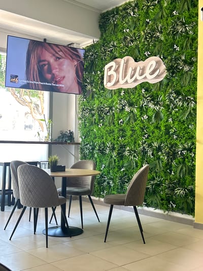 The image depicts the interior of a cafe or restaurant in Vera, Spain. A large, flat-screen television is mounted on the wall, displaying a close-up of a woman's face, with text overlay indicating "La Vida Es Una (From Puss in Boots: The Last Wish)" and the artist "KAROL G". To the right of the television, a wall is covered with artificial greenery, creating a lush, plant-covered backdrop. A light-colored, wooden sign with the word "Blue" in cutout letters is affixed to the greenery. In the foreground, a round, light-wood table is set with three upholstered chairs. The chairs are a neutral gray color with a quilted pattern and black legs. On the table, there are a few items, including a plastic cup and a small potted plant. The floor is tiled in a light, neutral color. A window is visible to the left of the television, showing a glimpse of trees and the outdoors, suggesting it is daytime.