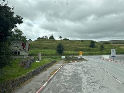 The image depicts an outdoor scene under a cloudy sky, likely in the High Peak region of the United Kingdom. In the foreground, a paved road curves to the right, bordered by a distinctive red and white striped kerb. To the left of the road, a narrow paved walkway runs alongside a low stone wall, backed by a grassy area.
Dominating the left side of the image is a large piece of industrial machinery, primarily grey with some red accents, possibly a crusher or similar equipment, resting on a concrete base. Beside it are several large, moss-covered rocks. Further back, a gentle, grassy hillside dotted with wildflowers and small trees stretches across the middle ground. A wooden fence runs along the top of a lower embankment.
Several signs are visible. A yellow triangular warning sign with a black arrow indicating a turn is present, accompanied by text below it. A white sign with "Regenera" is also visible. Further down the road, a sign with "20" inside a green circle indicates a speed limit, and below it, text reads "Twenty's Plenty Thank you for staying safe Please drive carefully". Another sign further in the distance appears to be a road sign.
The weather appears overcast, with heavy clouds dominating the sky, suggesting it might be a cool, damp day. There are no people visible in the image. The overall impression is one of a rural or semi-industrial landscape, possibly a lay-by or an area near a former industrial site, set against a backdrop of rolling hills.