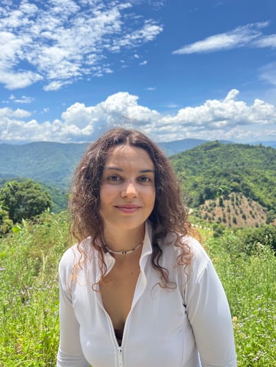 A medium shot captures a woman with curly brown hair and light skin, looking directly at the camera with a slight smile. She is wearing a white, zipped-up athletic top and a beaded necklace. She is positioned in the foreground with a lush, green mountain landscape and a bright blue sky with scattered white clouds in the background. The woman appears to be outdoors, possibly in Son Dien, Vietnam, given the verdant and hilly terrain suggestive of a rural or mountainous region.
