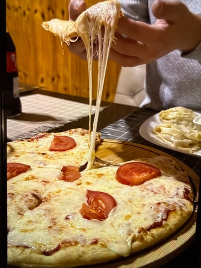 A close-up, eye-level shot captures a hand holding a slice of pizza, with long strands of melted cheese stretching downwards. The pizza is on a round wooden board, topped with melted cheese and slices of tomato. In the background, another plate of food, possibly dumplings, is visible. The environment appears to be indoors, with wooden paneling on the wall and a checkered tablecloth. The scene is likely in Artik, Armenia, based on the provided context.