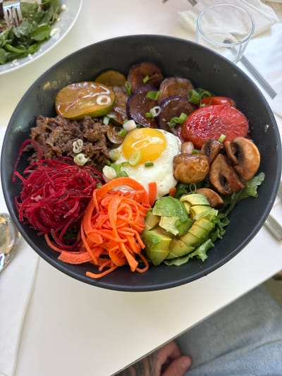 A black bowl of bibimbap is centered on a white table. The bibimbap contains a fried egg, seasoned beef, sliced eggplant, sliced tomatoes, mushrooms, shredded carrots, shredded beets, and avocado. The bowl is placed next to a partially visible salad in a white bowl and a clear glass. The background shows a white napkin and a metallic utensil. The scene is lit by overhead lighting, casting soft shadows. The overall impression is a well-prepared and visually appealing meal. The location is a restaurant in Burnaby, Canada.