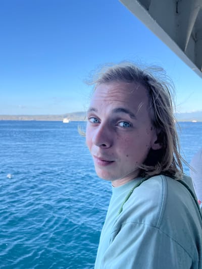 A young Caucasian man with blonde hair and blue eyes is leaning towards the camera, looking directly at it. He is wearing a light teal t-shirt and has a light scattering of freckles across his nose and cheeks. The background is a bright blue sky and the shimmering blue water of the ocean. In the distance, a hazy coastline with some greenery is visible, along with a white ship. The perspective suggests the man is on a boat, with a portion of the boat's structure visible at the top right of the image. The overall lighting is bright and sunny, indicating daytime. The image is a close-up portrait, with the man's face occupying a significant portion of the frame. The location is the ocean near the city of Ketapang, Indonesia.