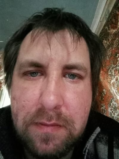 A close-up shot of a middle-aged man's face. He has fair skin, blue eyes, and dark hair with some graying. He has a beard and mustache. His face shows signs of acne scarring. He is wearing a dark-colored, possibly wool, sweater. The background includes a pale green wall and a patterned carpet or tapestry, suggesting an interior setting, likely a home. The lack of external cues prevents confirming that the photograph was taken in Parabel, Russia. There is no interaction or action evident in the photo, it is a static portrait.