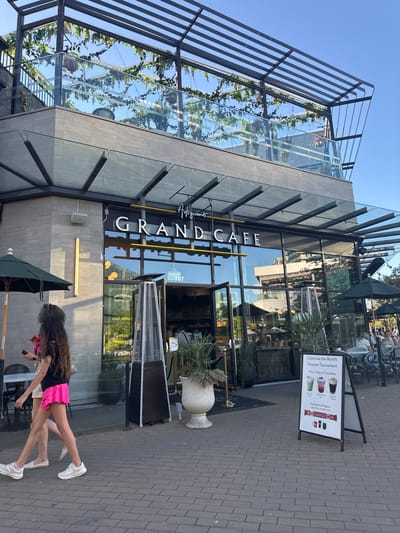 The image depicts the exterior of the Grand Cafe, a restaurant located in West Vancouver, Canada. Two young females are walking on a paved walkway in the foreground, with one wearing a black t-shirt and a bright pink skirt, and the other in a white top and a tan skirt. They are walking past a large, modern building with a glass facade and an outdoor seating area. The Grand Cafe's sign is prominently displayed above the entrance. Several outdoor tables with umbrellas and chairs are visible on the patio, with some patrons seated at them. A sign board in front of the cafe advertises "Celebrate the World's Greatest Tournament With a Taste of Canadiana". The overall environment suggests a sunny day in an urban setting.