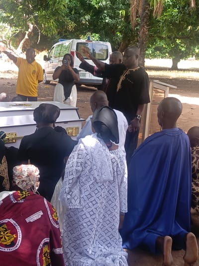 A funeral service is taking place in what appears to be Ninzo South, Nigeria. The scene includes a white coffin with gold accents, around which mourners are gathered. Some mourners are kneeling, while others are standing. Individuals are wearing a variety of clothing including white lace, black attire with gold embroidery, yellow shirts, and blue fabric. A person is taking a photo with a phone. An ambulance is parked in the background. The scene is under trees and in an outdoor environment. A plate on the car says "06C-07KD".