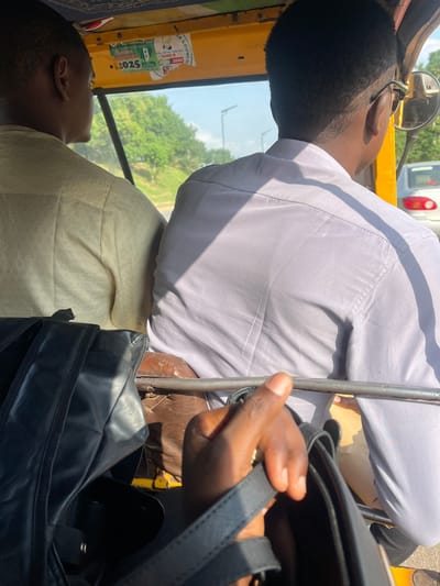 The image shows two individuals inside a vehicle, likely a rickshaw or a similar open-air transport, as indicated by the visible yellow exterior and a mirror. The scene appears to be outdoors during the daytime, with bright sunlight casting shadows and highlighting textures. The overall atmosphere suggests a journey or commute.
In the foreground, a person's hand is visible, holding onto a black bag. The hand has dark skin tone. The black bag is open, revealing a dark interior. Behind this, a person wearing a light-colored, possibly white or light grey, long-sleeved shirt is seated. The shirt is slightly wrinkled, and sunlight illuminates its fabric. The person's back and side are visible, with the head turned away from the camera.
In the background, another person is seated, with their head leaning back and eyes closed, suggesting rest or contemplation. This person has dark skin and short hair. They are wearing a light-colored garment, possibly the same as the person in the foreground or a cream-colored patterned fabric. The interior of the vehicle is visible, including the yellow framing and parts of the seats.
Through the window or opening of the vehicle, lush green trees and foliage are visible, indicating a possibly rural or semi-urban setting. The sky is bright, suggesting good weather.
On the side of the vehicle, some stickers are affixed. One sticker appears to be an election-related campaign poster, with text such as "COUNCIL B," "ZONE B," and "2025." Another sticker has text that is partially obscured but includes what might be "NIGERIA 2025." The location context of Galadimawa Village, Nigeria, aligns with the presence of this type of transport and the visual cues of the environment. No specific emotions are clearly discernible from the individuals, but the person with eyes closed might be experiencing a sense of relaxation.