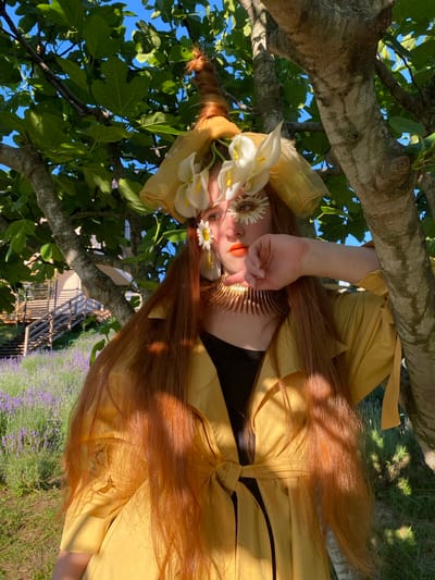 This media file shows a person with long, auburn hair wearing a yellow garment, possibly a dress or coat, and a headdress adorned with white calla lilies and small daisies. A golden comb-like hair accessory is visible near their forehead. The person's face is partially obscured by their hand, which is touching their cheek. They are lying or leaning against a tree with thick branches and green leaves, with the sky visible in the background. A field of lavender and a wooden structure can be seen in the distance. The lighting suggests it is daytime, with sunlight casting shadows. The overall mood is serene and ethereal. The location is identified as Verkhneye Uchdere, Russia.