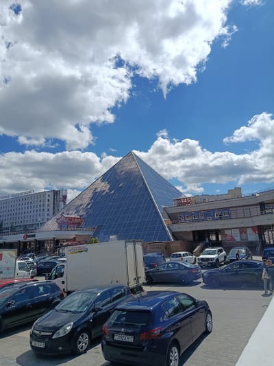 This image captures a daytime scene in Vitsebsk, Belarus, under a bright, partly cloudy sky. The focal point is a modern glass pyramid building, likely a commercial center or office complex, with its facets reflecting the blue sky and white clouds. To the left of the pyramid stands a large, white multi-story building with numerous windows, displaying Cyrillic text that appears to be a hotel name.
In the foreground, a parking lot is filled with various cars and a large white truck. Cars are parked in rows, and some appear to be driving or maneuvering within the lot. The atmosphere seems bustling, suggesting a typical day in a commercial area.
Visible text includes "ОДНАЯ ВІЦЕБШЧЫНА" and "STEPWAY" on a car. Another car has the text "PEUGEOT 3192KH-2", and a truck has Cyrillic text that includes "PEMOHT" and a phone number. Additionally, advertisements with discounts like "-70%" are visible on the commercial building. The presence of multiple vehicles and the architectural style suggest an urban setting. The overall impression is one of everyday activity in a modern city.