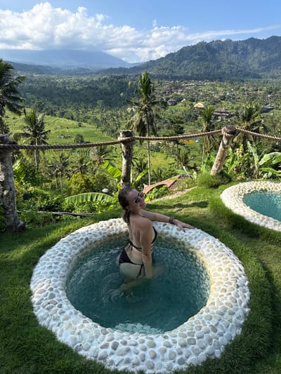 The image depicts a woman in a black bikini relaxing in a small, circular pool filled with clear turquoise water. The pool is surrounded by lush green grass and a decorative rim of smooth, light-colored stones. The woman is smiling and wearing sunglasses, with her left arm resting on the edge of the pool. Behind her, a stunning panoramic view of the Sidemen Valley in Bali, Indonesia, unfolds. The valley is characterized by vibrant green rice terraces cascading down the hillsides, dotted with small villages and palm trees. In the distance, a majestic mountain range rises, partially shrouded in a hazy sky with scattered clouds. The scene conveys a sense of tranquility and natural beauty, highlighting the serene environment of Sidemen, Indonesia.