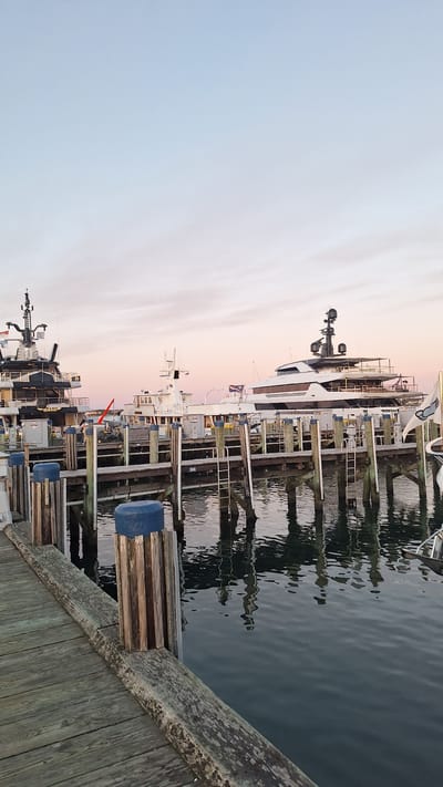 The image depicts a serene harbor scene in Nantucket, United States of America, at dusk. In the foreground, weathered wooden planks form a pier, leading the eye towards the water. Several mooring posts, some topped with blue caps, punctuate the pier's edge. Beyond, a fleet of luxurious yachts is docked.
The most prominent vessel on the left is a black and white yacht with a tall mast. To its right, a smaller white boat with a tower stands. Further to the right, a large, sleek white yacht dominates the scene, its modern design contrasting with the rustic pier. The yachts are moored to a wooden dock structure.
The water in the harbor is calm, reflecting the structures and the soft hues of the twilight sky. The sky itself transitions from a pale blue at the top to a gentle pink and orange near the horizon, indicative of a sunset. The overall atmosphere is one of tranquility and maritime elegance. There are no people visible in the image, emphasizing the quiet stillness of the harbor at this time of day. The lighting suggests late afternoon or early evening.