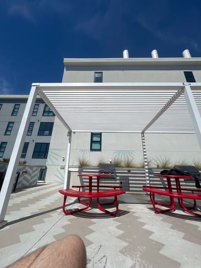 This image depicts a sunlit outdoor communal patio area in Gainesville, United States of America, likely on a rooftop or elevated terrace, under a clear blue sky.

In the mid-ground, a white metal pergola structure, composed of numerous parallel slats, extends across the scene, casting distinct striped shadows onto the light-colored concrete ground and the adjacent building wall. Beneath the pergola are two bright red, metal picnic-style tables, each with attached curved benches. The table on the left is partially visible in the foreground, while the table on the right has a dark garment, possibly a black shirt or jacket, draped over one of its benches.

Between the two red tables, against the backdrop of a light grey building wall, are two built-in outdoor barbecue grills with corrugated metal covers. Above these grills, a planter box contains sparse, light green ornamental grasses. The building wall itself features decorative stencil patterns resembling palm fronds in a lighter shade of grey. An "EXIT" sign is visible on this wall to the left of the grills.

Further back, the light grey buildings rise multiple stories, displaying rectangular dark-framed windows. The roofline of the closer building features three white, cylindrical ventilation units. Another building section to the far left exhibits a diagonal zig-zag pattern in light blue and white on its lower exterior. The ground is paved with light-colored concrete featuring a repeating zig-zag pattern in a slightly darker grey.

In the lower left foreground, a portion of a person's bare legs, specifically the upper thighs and knees, is visible, indicating the perspective of someone seated or reclining on the patio. No other individuals are present in the frame. The scene is brightly illuminated by direct sunlight, creating sharp shadows.