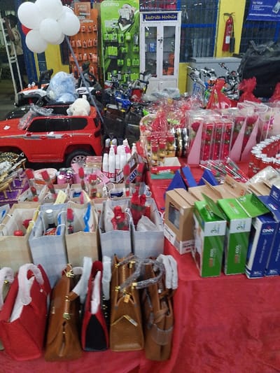 The image shows a table laden with gifts and merchandise. There are several purses, gift bags with items like roses and snacks, and various packaged goods. In the background, there's a small red toy car, bunches of white balloons, and other items such as bicycles and shelving units with products. A fire extinguisher is visible on the wall, suggesting a retail or store environment.