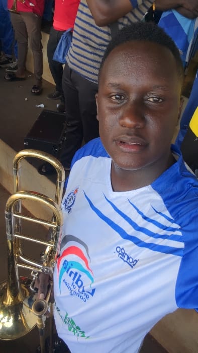 A man is seen wearing a blue and white jersey with sponsor logos. He is partially visible, as he is laying down, angled towards the camera. A gold trombone is positioned above him, possibly being held by another person whose hand is barely visible. Other individuals, some with bags, are seen lying down nearby. The scene appears relaxed and informal.