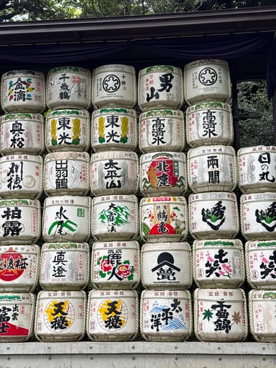 This image captures a vibrant display of traditional Japanese sake barrels, known as *kagami-biraki* barrels or *komodaru*, stacked neatly in horizontal rows. There are at least five visible rows and eight columns of barrels, filling the majority of the frame.
Each barrel is cylindrical, wrapped in a straw mat (komo) and secured with rope, featuring a distinct paper label (noren) on its front. These labels showcase a rich variety of calligraphic brand names, traditional Japanese art, logos, and crests, often in black ink with highlights of red, green, blue, yellow, and pink. Many labels also include the term "清酒" (Seishu - refined sake) and "登録商標" (Tōroku Shōhyō - Registered Trademark).
Notable examples of visible text and imagery include "上川大雪" (Kamikawa Taisetsu) from Hokkaido with a circular crest, "男山" (Otokoyama), "高清水" (Takashimizu), "千歳鶴" (Chitose Tsuru), "白鶴" (Hakutsuru), "真澄" (Masumi), "越後桜" (Echigozakura) from Niigata, and "島根" (Shimane), among many others, representing various breweries across Japan. Some barrels display intricate designs like cranes, cherry blossoms, peonies, dragons, or stylized kanji characters. English text such as "KAMIKAWA TAISETSU HOKKAIDO" and "ILAKA JAPANESE SAKE ATELIER" is also present on some labels.
The barrels are displayed outdoors, possibly under a covered structure or an overhang, indicated by the dark, draped fabric and a structural beam on the left, and a concrete divider on the right. Green foliage from trees is visible in the background on the left, suggesting a natural setting. The lighting is soft and even, indicating daytime, possibly overcast conditions or shade. This arrangement is typical of sake offerings displayed at Shinto shrines or temples, often seen at locations like Meiji Jingu in Shibuya, Japan, to honor deities and celebrate the breweries. The display evokes a sense of tradition and cultural heritage.
