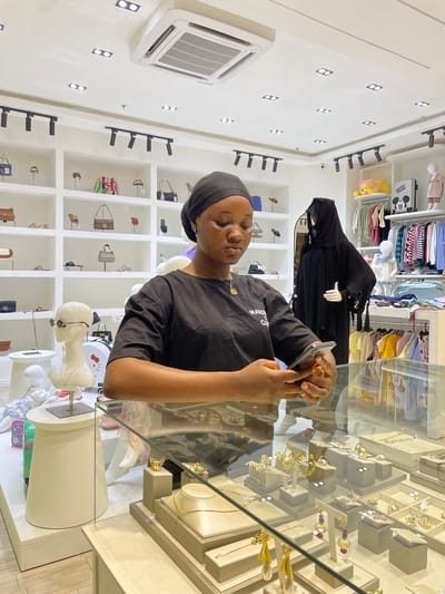 In a brightly lit boutique in Unguwar Sarki, Nigeria, a young Black woman wearing a black headscarf and black t-shirt is focused on her smartphone, holding it in both hands. She is positioned in the foreground, partially obscuring a glass display case filled with a variety of gold jewelry, including necklaces, rings, and earrings. Behind her, shelves are stocked with handbags and hats, and to her right, mannequins are dressed in children's clothing and traditional Islamic attire. The store's ceiling features modern recessed lighting and an air conditioning unit. The overall environment suggests a retail space specializing in fashion accessories and clothing.