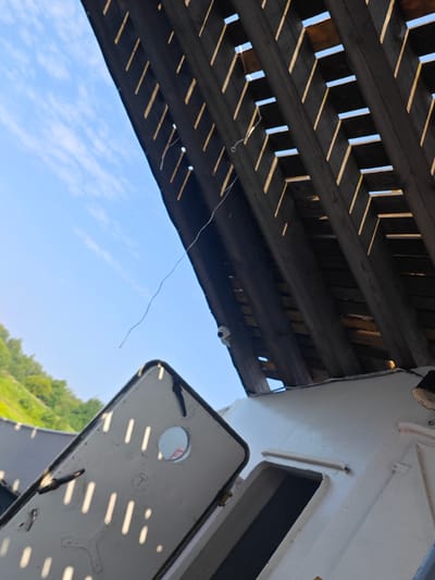 An exterior shot taken from a low angle, looking up towards a wooden structure with a slatted roof, allowing sunlight to filter through. In the foreground, a white, rectangular object with a circular opening and a metallic trim is propped open, casting striped shadows from the sunlight. A thin white wire hangs from the wooden structure towards the white object. To the left, a green, tree-covered hillside is visible under a bright blue sky with scattered white clouds. The scene appears to be outdoors in a rural or coastal setting, possibly near Novonazimovo, Russia, given the natural elements and the style of the structure.