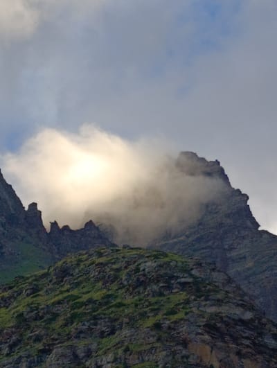 The image depicts a dramatic mountain landscape in Damphug, India. The foreground features a steep, rocky slope covered in sparse green vegetation. Jagged, dark grey peaks rise sharply from this slope, with some of their formations resembling jagged teeth. A thick bank of luminous white clouds is partially obscuring the upper portions of these mountains, with sunlight breaking through the clouds to create a bright, glowing effect. The sky above is a muted blue, with lighter clouds scattered throughout. The overall scene conveys a sense of rugged, natural beauty and the atmospheric conditions of a mountainous region.