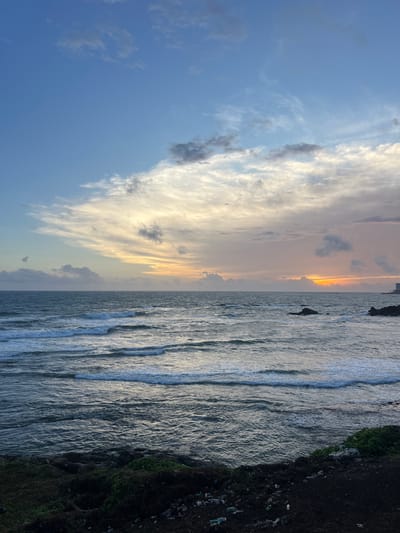 This image captures a scenic view of the ocean at dusk, likely in Galle, Sri Lanka. The scene is divided into two distinct halves. The left side of the image is dominated by a vast expanse of sky filled with dramatic clouds. The clouds are illuminated by the setting sun, creating a gradient of colors from a deep blue on the left to warm hues of orange and yellow closer to the horizon. Wispy, elongated clouds streak across the sky, catching the light and adding texture to the scene.
The right side of the image features the ocean, with waves gently rolling towards the shore. The water appears to be a deep blue-grey, reflecting the muted light of dusk. White foam marks the crests of the waves, creating a dynamic contrast with the darker water. Along the far right edge of the image, a dark, rocky embankment is visible, covered in patches of green vegetation. This embankment is noticeably littered with various pieces of plastic waste, suggesting a human impact on the natural environment.
The time of day is clearly indicated by the sunset colors in the sky and the dimming light over the ocean. The weather appears to be clear, with only scattered clouds. The presence of the ocean, sky, and rocky shoreline strongly points to a coastal location. The emotion evoked by the scene is a mix of natural beauty and a touch of melancholy due to the visible pollution. No text is visible in the image.