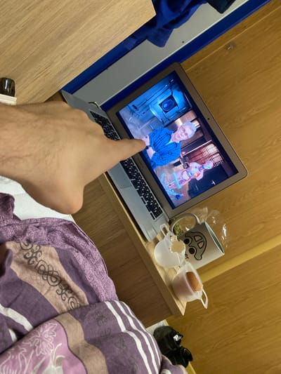 The image shows a person's hand pointing at a laptop screen. The laptop is sitting on a light brown wooden surface next to a bed covered in a purple and white striped blanket. On the screen, a man wearing a blue checkered shirt is visible next to a human anatomy model. Next to the laptop is a mug with a car design, an egg in a plastic cup, and broken eggshells in a smaller plastic cup. Two black shoes are visible in the background. The room has light brown wooden walls. The location of the image is likely in Koskolovo, Russia.