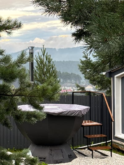This is a vertical, eye-level, outdoor shot of a balcony in Kusa, Russia. The foreground is dominated by the needles of pine trees, some of which are laden with water droplets, suggesting recent rain or mist. Beyond the trees, a dark, modern balcony railing encloses a space where a large, dark, rounded object, possibly a covered hot tub or an outdoor grill, is partially shrouded in a light-colored fabric cover. To the right of this object, a simple wooden bench with a metal frame is visible.
The background opens up to a hazy, green landscape of rolling hills and a small cluster of houses, hinting at a village or town nestled in the valley. The sky is overcast with soft, diffused light, indicating either early morning or late afternoon, with a touch of yellow in the clouds suggesting the sun may be trying to break through. The weather appears damp and cool.
The overall mood is one of quiet observation, with the natural elements of the pine trees and the distant landscape contrasting with the man-made structures of the balcony and the covered object. There is no visible text in the image.