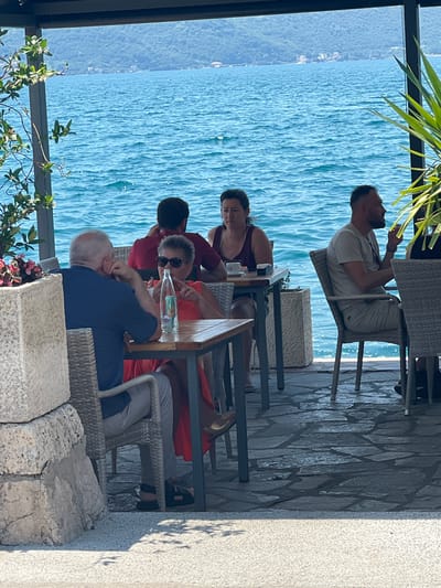 This is a horizontal, medium shot of a waterside restaurant in Risan, Montenegro. Four people are seated at tables. In the foreground, a man with gray hair and a blue shirt is sitting at a table with a woman wearing an orange dress. The woman is wearing sunglasses and looking at the man, who has his hand to his ear. A bottle of water and two glasses are on the table. In the background, a man in a red shirt sits at another table, and a woman in a dark red top is seated further back. The restaurant is open-air, with a partial roof and a view of the Bay of Kotor. The water is a deep blue, and hills with trees and buildings can be seen in the distance. The ground is paved with irregular stone tiles.