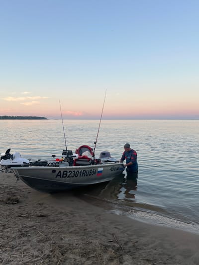 In a sideways shot, a person in a red and blue long-sleeved shirt and a grey beanie is partially submerged in calm, rippling water next to a grey fishing boat. The boat is beached on a sandy shore, with its bow pointing towards the viewer. The boat is equipped with two fishing rods, one extending from the front and another from the middle, both with red tips. A white electric trolling motor is attached to the back of the boat. The boat also displays the registration number "AB2301RUS54" in large black lettering. A Russian flag sticker is visible on the stern of the boat.

The scene depicts a tranquil sunset over a body of water. The sky transitions from a pale blue at the top to soft shades of pink and orange closer to the horizon, indicating either sunrise or sunset. The water reflects the colors of the sky, creating a peaceful ambiance. The sandy shore is visible in the foreground, with a few dry twigs scattered on it. The overall mood is one of quiet reflection and enjoyment of nature. The person appears to be adjusting something on the boat. The location context is Borovoye, Russia.