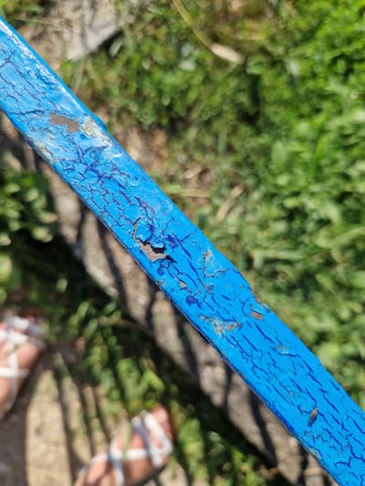The image shows a close-up of a weathered, bright blue painted metal bar, likely part of a railing or fence, set against a blurred background of green grass and some concrete. The paint on the bar is cracked and peeling, revealing rust underneath in several areas. The texture of the paint is rough and uneven. In the lower left and lower right corners of the image, parts of two pairs of feet wearing white sandals are visible. The feet appear to be standing on a rough, possibly sandy or gravelly surface, with shadows cast across it. The overall impression is an outdoor, possibly urban or park-like setting, given the presence of the railing, grass, and feet. No specific location in the city of German, Bulgaria can be determined from the image.