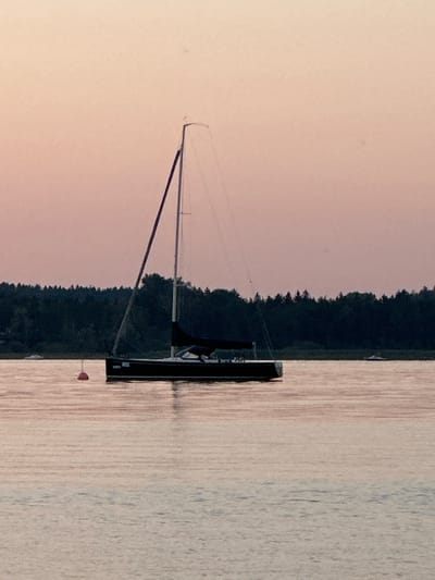 The image depicts a sailboat on a body of water, likely a lake or bay, at dusk. The sky is a soft, peachy-pink, transitioning to a lighter hue towards the horizon. A dark green treeline lines the background, suggesting a forested shore. The water is calm, reflecting the pastel colors of the sky and the silhouette of the boat.
The sailboat is dark-colored and has a single mast with rigging visible. A dark sail is partially furled. A small, red buoy is visible in the water to the left of the sailboat. In the distance, another small white boat can be seen near the treeline. There are no people visible in the image. The scene appears tranquil and serene.
While the image itself does not provide definitive clues to its location, the description requests a potential location in Prien am Chiemsee, Germany. Prien am Chiemsee is situated on the shore of Lake Chiemsee, a large Bavarian lake known for its scenic beauty and sailing opportunities. The presence of a sailboat, treelined shores, and calm waters aligns with the characteristics of Lake Chiemsee. However, without further contextual information or identifiable landmarks, this specific location remains speculative based on the provided image.