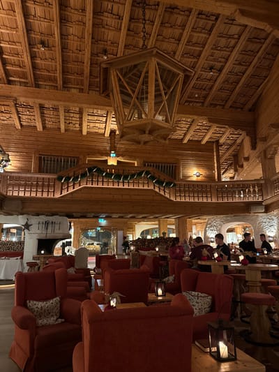 A cozy, rustic interior features warm lighting and wooden architecture. Ornate wooden ceilings and a large, cage-like chandelier dominate the upper space. Below, plush red armchairs are arranged in rows, facing towards a fireplace and a snow-decorated area. People are seated at tables in the background, creating a lively atmosphere. A staircase with garland runs along the side.