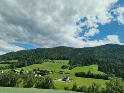 A broad, sunlit, rural landscape scene, typical of Scheifling, Austria, under a partly cloudy sky. Rolling green hills, covered in lush grass and scattered trees, dominate the foreground and mid-ground. In the middle distance, a small cluster of houses, with pitched roofs and some featuring solar panels, are nestled amongst the greenery. A dense, dark green coniferous forest blankets the steeper slopes of a large mountain that rises in the background. The sky is a mix of bright blue patches and dramatic, grey, textured clouds. The overall impression is one of a peaceful, serene, and natural environment.