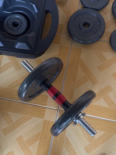 The image shows a dumbbell with two weight plates attached to it, lying on a tiled floor. The dumbbell has a black grip with red bands around the middle and a silver threaded end where the weight plates are attached. The weight plates are dark gray with a textured surface and a central hole. One of the plates has visible text that appears to read "2.5 KG". To the left of the dumbbell is a black plastic case, likely for storing dumbbells, with a weight plate nestled inside. The case has a handle and a contoured interior. The floor is tiled with a pattern of light and dark beige rectangular tiles arranged in a parquet style. The lighting suggests an indoor setting, possibly a home gym or workout area. No people are visible in the image. The scene is still and focused on the weightlifting equipment.