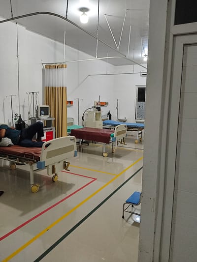 The image depicts a hospital ward with several beds. In the foreground, a person is lying on a bed, appearing to be asleep or unconscious, with their head resting on a pillow. The bed is equipped with medical equipment, including an IV pole and a monitor. The room is brightly lit by overhead fluorescent lights. The floor is tiled and marked with colored lines, possibly indicating different zones or pathways. The background shows more hospital beds, medical equipment, and a curtain. The overall impression is that of a functional but somewhat stark medical facility. The location is identified as Meunasahaluedrienlhoksukon Barat, Indonesia.