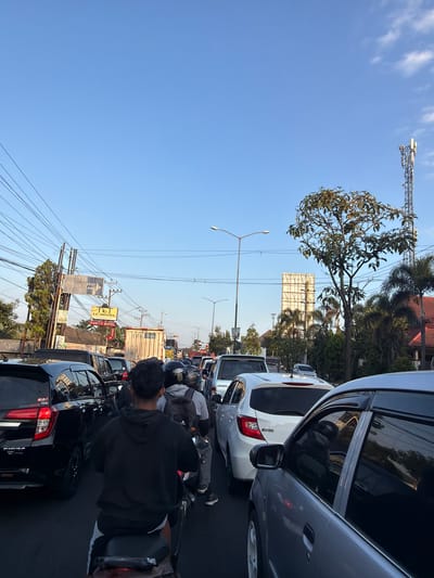 A street scene in Danurejo, Indonesia shows a traffic jam with many vehicles on the road, including cars, motorcycles, and a truck. The sky is clear and blue. Power lines crisscross overhead. A cell tower is visible in the distance. There are trees and buildings on either side of the street. A person on a motorcycle is in the foreground, facing away from the camera. The traffic appears to be moving slowly.