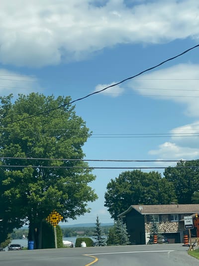 This is a ground-level, eye-level, static, medium shot photograph taken in East Hawkesbury, Canada, on a sunny day with scattered clouds. The image captures a residential street scene bordering a body of water. In the foreground, a paved road curves to the left. A large deciduous tree with lush green foliage dominates the left side of the frame, partially obscuring a blue trash bin and a parked silver sedan. To the right of the tree, a diamond-shaped traffic sign with a black and yellow checkered pattern and a black arrow pointing left is mounted on a pole.
Further into the scene, a two-story house with a stone and dark siding exterior is visible. The house has a dark-colored garage door and several windows. In front of the house, small evergreen trees with orange striped bases are planted. A white propane tank sits on the right side of the yard. A boat is moored near the water's edge.
Beyond the house and trees, a wide expanse of water is visible, with a distant shoreline featuring more trees and buildings. Power lines stretch across the sky, intersecting the scene. A sign indicating "CONSTRUCTION ZONE BEGINS" with orange traffic cones is present near the right edge of the frame, suggesting ongoing road work. The overall impression is one of a peaceful, semi-rural residential area.