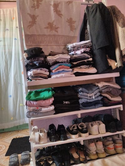 An indoor scene in Witeithie ward, Kenya, depicts a three-tiered white wooden shelving unit organized for clothing and footwear storage.
On the topmost rail, a light-colored blanket or towel featuring repeating cherub motifs in brown hangs draped. To its right, dark garments are suspended from hangers, with another lighter textured item partially visible.
The top shelf contains three distinct stacks of folded apparel: a mixed dark and light stack on the left, a predominantly light-colored stack in the center including white, blue, and pink items, and a mixed dark and light stack on the right.
The middle shelf also holds three stacks: a green garment atop a pink and a grey item (the grey item bearing "MERSHIER" text) on the left, dark-colored items in the center (one with a spider-like emblem), and folded blue denim garments atop other items on the right.
The bottom shelf is dedicated to footwear, displaying from left to right: two pairs of white sneakers, two pairs of black platform shoes, two pairs of dark-colored low-top sneakers, and two pairs of dark clogs.
On the tiled floor in front of the unit, two pairs of dark grey Crocs-style clogs are present. Further pairs of shoes, including black flats, heels, and multi-colored sneakers, are visible on the floor beneath and around the base of the shelving unit.
The background features a translucent white curtain adorned with small, colorful shapes on the left. The wall behind the unit is a light, possibly lavender hue. The floor is tiled with a light brown pattern, and a small black mat rests on the tiles.