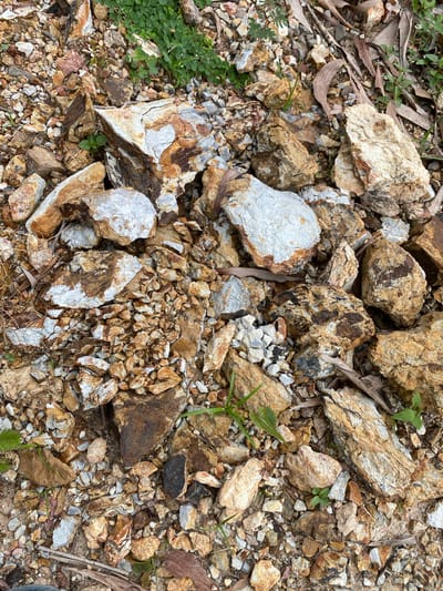 The image depicts a close-up, overhead view of a pile of rocks and gravel. The rocks are varied in size, shape, and color, ranging from light tan and white to dark brown and reddish-brown. Some rocks show white mineral deposits. Small green plants are interspersed among the rocks, and dry, fallen leaves are also visible. The scene appears to be an outdoor environment, possibly a construction site or a natural rocky area. There are no people or animals visible in the image. The location is identified as Ko Siboya, Thailand.