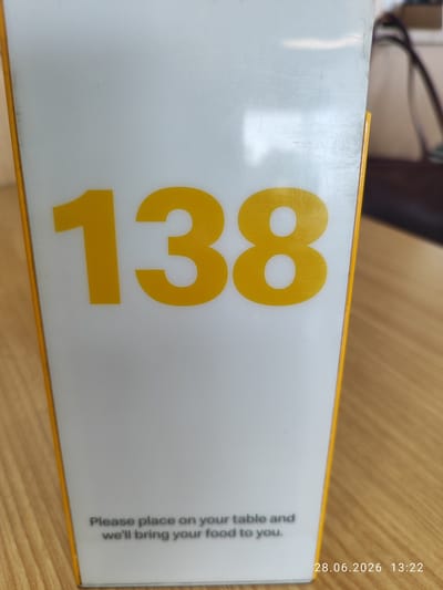 A close-up shot of a white table number sign with yellow digits "138" is shown. Below the number, a message in gray text reads, "Please place on your table and we'll bring your food to you." In the bottom right corner, a date stamp reads "28.06.2026 13:22". The sign is positioned on a wooden surface, likely a table. The background shows a blurry interior environment. The image is taken in Northampton, United Kingdom.