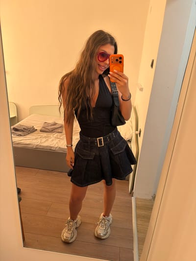 A young woman with long, wavy brown hair stands in front of a mirror, taking a selfie. She is wearing a black halter top, a dark denim skirt with a belt, and white sneakers. She has on large pink-tinted sunglasses. Her right hand holds an orange phone case with a black phone. A black purse hangs from her left shoulder. In the background, a bed with gray linens is visible in a room with light beige walls and wooden flooring. The scene appears to be indoors, possibly in a hotel room or apartment, in the city of Svishtov, Bulgaria.