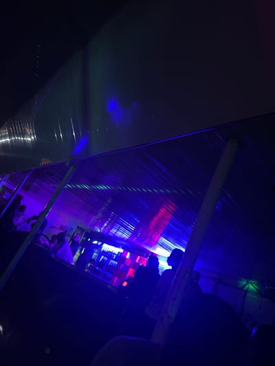 The image depicts an indoor scene at a bar or lounge in Wumat, Nigeria, bathed in vibrant blue and purple lighting, with streaks of red and green light reflecting off the corrugated ceiling. Silhouetted figures are visible, some standing near a bar that appears stocked with bottles. The overall atmosphere suggests a lively, nighttime setting, possibly a nightclub or entertainment venue.