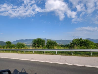 The image depicts a roadside view on a clear day with a blue sky filled with scattered white clouds. In the foreground, a two-lane asphalt road is visible, with a white line marking the lane separation. A metal guardrail runs along the side of the road, separating it from a grassy embankment. Beyond the embankment, a line of green trees and bushes forms a natural barrier. In the background, rolling hills and mountains are covered in vegetation, suggesting a rural or mountainous landscape. The scene appears to be captured from a moving vehicle, as indicated by the slight blur and the reflection of what looks like a car mirror and shadows on the road. There is no visible indication of the city of Dzhurovo, Bulgaria, within the frame.