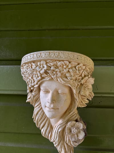 A cream-colored, weathered wall planter, sculpted as the head and upper torso of a female figure, is mounted centrally against a dark olive-green wooden plank wall. The figure faces forward with eyes closed, a subtle smile, and long, wavy hair adorned with a large flower on its right side. A detailed floral wreath forms a crown around her head, topped by an ornate decorative band, collectively serving as the planter's rim. Traces of dirt, discoloration, and fine cobwebs are visible on the sculpture, particularly within the crevices of the hair and flowers, and on the decorative band. The background consists of horizontally aligned, painted wooden planks exhibiting some wear and faint cobweb presence on the left side. The scene is located in the city of Stiperstones, United Kingdom.