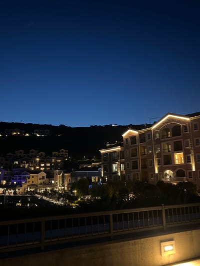 The image depicts a nighttime view of a coastal town, specifically the area around the Porto Montenegro marina in Gošići, Montenegro. The foreground features a dark, textured concrete wall with a rectangular, illuminated light fixture. Above this wall is a dark metal railing. Beyond the railing, the scene opens to a cluster of buildings illuminated by warm artificial lights.
The buildings are multi-story structures with architectural details like arched windows, balconies, and decorative trim. Some buildings are adorned with subtle exterior lighting, highlighting their features. The architectural style appears to be a blend of Mediterranean and modern influences.
Further in the background, more buildings are visible, cascading up a dark, hilly landscape. These structures also feature illuminated windows, suggesting occupancy. The overall impression is of a well-lit, developed waterfront community at dusk or early night. The sky is a deep, clear blue, transitioning to black at the top of the frame, indicating either late evening or early morning. A faint streak, possibly a contrail from an aircraft, is visible in the upper portion of the sky. The scene is tranquil and suggests a populated but quiet atmosphere.
