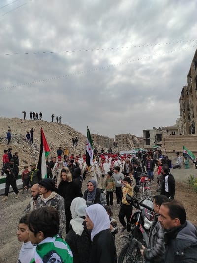 The image depicts an outdoor procession taking place in Ash-Shaghour Municipality, Syria, under an overcast sky. In the foreground, a diverse crowd of individuals, including men, women, and children, occupies a dirt and gravel path. A Palestinian flag (black, white, green, red stripes) is held upright on the left, next to a Syrian opposition flag (green, white, black with three red stars).

In the midground, a larger procession of people, many dressed in white garments with red head coverings, proceeds along the path. Several additional Syrian opposition flags are visible within this marching group. A young male in a yellow top is observed holding a mobile phone in a recording posture. A black motorcycle is parked among the individuals in the midground.

On the far left background, a substantial mound of construction debris is visible, with approximately ten dark-clad figures standing atop it, observing the scene. To the right background, multiple multi-story residential buildings exhibit extensive structural damage, including exposed interiors, missing walls, and collapsed sections. Thin black lines, likely power or communication cables, traverse the cloudy grey sky. The environment suggests an urban area severely affected by destruction.