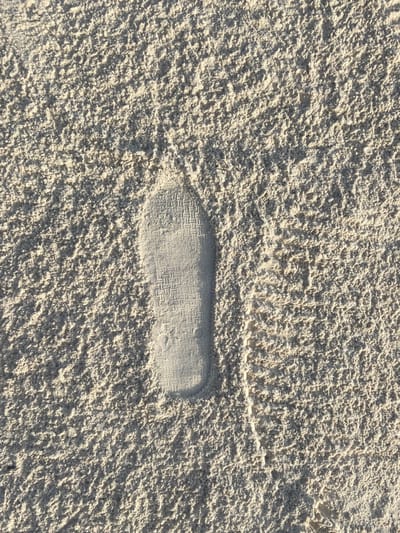 This media file presents a close-up, overhead view of a single, well-defined footprint impressed into light-colored sand. The sand occupies the entire frame, displaying a rough, uneven texture composed of countless small grains, creating numerous highlights and shadows. The footprint is centrally located, showing the distinct outline and flat impression of a shoe sole. Inside the compressed area of the print, a clear and intricate geometric tread pattern is visible, resembling a lattice or woven design. The strong, natural lighting, likely from a bright sun, casts sharp, contrasting shadows that enhance the depth and texture of both the undisturbed sand and the footprint. The scene suggests a serene, natural environment, consistent with a beach. Given the location context of Kiwengwa, Tanzania, this image captures a detail from a sandy shore during daytime, under clear and sunny conditions. No individuals or other objects are visible, and no literal text is present beyond the shoe's sole pattern.