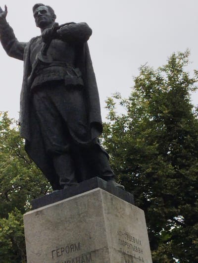 A low-angle, full shot captures a dark, weathered statue of a man in uniform, possibly a soldier or a military leader, standing proudly on a concrete pedestal. The statue is depicted from the waist up, with the figure's right arm raised in a gesture of salute or command, and a hand clutching a baton or rifle. The man wears a coat draped over his shoulders and a belt cinched around his waist. The statue is positioned outdoors, with lush green trees forming a dense backdrop. The sky above is a pale, overcast gray, suggesting a cloudy day.

Visible text on the pedestal reads in Russian: "ГЕРОЯМ" (To the Heroes) followed by what appears to be names and possibly dates, though some are obscured or difficult to discern. Further down, another line reads "КОНОНОВУ МХ" (Kononov M.Kh.) and "ЛАВРЕНТЬЕВУ В.Н." (Lavrentyev V.N.). This monument is located in Kingisepp, Russia. The overall mood is solemn and commemorative, honoring significant figures from history.