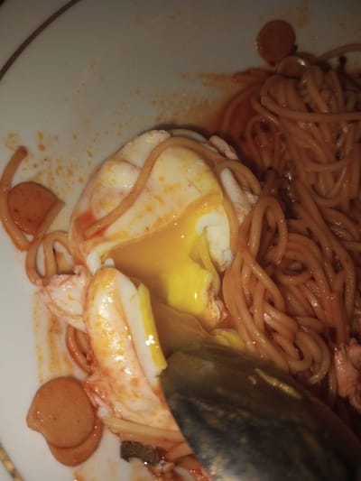 A close-up, high-angle view depicts a meal served on a white ceramic plate with a subtle gold-colored rim. The dish consists of spaghetti or thin noodles coated in a reddish-orange sauce, numerous circular slices of reddish-brown hot dogs or sausages, and a central, partially disrupted egg. The egg, likely poached or soft-boiled, shows a vibrant yellow, liquid yolk actively spilling from its broken white and mingling with the surrounding sauce and noodles. A dark, metallic spoon is partially visible in the lower right, submerged in the food, suggesting an ongoing act of consumption. Splatters of the reddish-orange sauce are present on the plate's rim. The specific location within Bagochen Boolingpang Ueling, Bhutan, is not discernible from the visual evidence presented in the image.