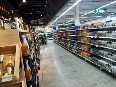 The image depicts the interior of a supermarket in El Valle del Espíritu Santo, Venezuela. The aisle features shelves stocked with various beverages, including bottled water, juices in cartons, and alcoholic drinks. The lighting is bright, suggesting a well-lit commercial environment. In the foreground, on the left, a wooden display holds several bottles of wine, one of which has a price tag of 18.66. To the right, a long aisle stretches into the distance, lined with shelves on both sides packed with a variety of bottled and boxed drinks. A sign indicating "7 PASILLO" (7 AISLE) is visible hanging from the ceiling. The floor is tiled with a light-colored speckled pattern. No people are clearly visible in the image.