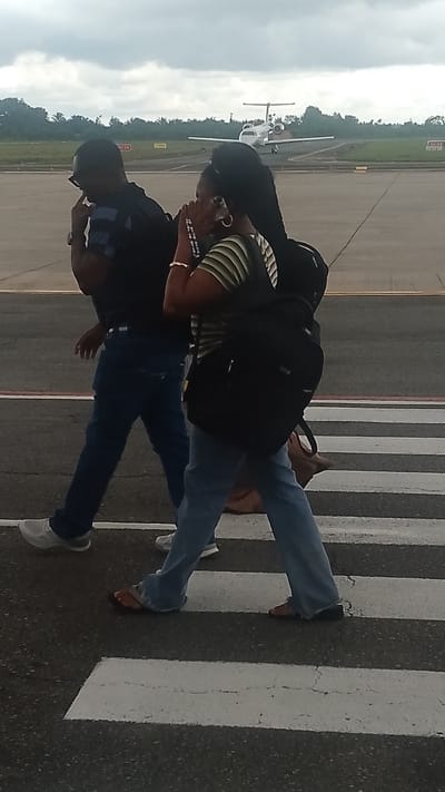 The image captures two individuals walking across a pedestrian crossing on an airport tarmac. A man in a dark blue collared shirt and jeans, wearing sunglasses and a watch, is on the left. Beside him, a woman in a striped shirt and light blue jeans is talking on her phone and carrying a black backpack. She also has a light brown bag slung over her shoulder.
In the background, a white private jet is visible on the runway, suggesting an active airport environment. The sky is overcast, indicating a cloudy day. The ground is asphalt, marked with white lines for the pedestrian crossing. The presence of the aircraft and the tarmac clearly establish the setting as an airport. The individuals appear to be navigating the airport, possibly en route to or from a flight. The overall mood is neutral, focusing on the action of walking and the airport environment. There is no visible text in the image.