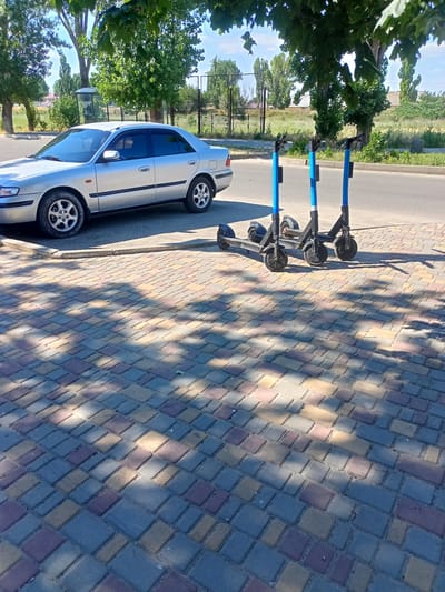 A silver sedan is parked on the side of a street. Three blue electric scooters are parked next to the car on the sidewalk. The sidewalk is made of interlocking pavers. Trees with green leaves are in the background. The sky is blue. This image was taken in Bilhorod-Dnistrovskyi, Ukraine.
